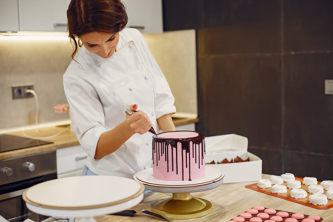 Smiling young woman squeezing chocolate from pastry bag on cake in pastry store